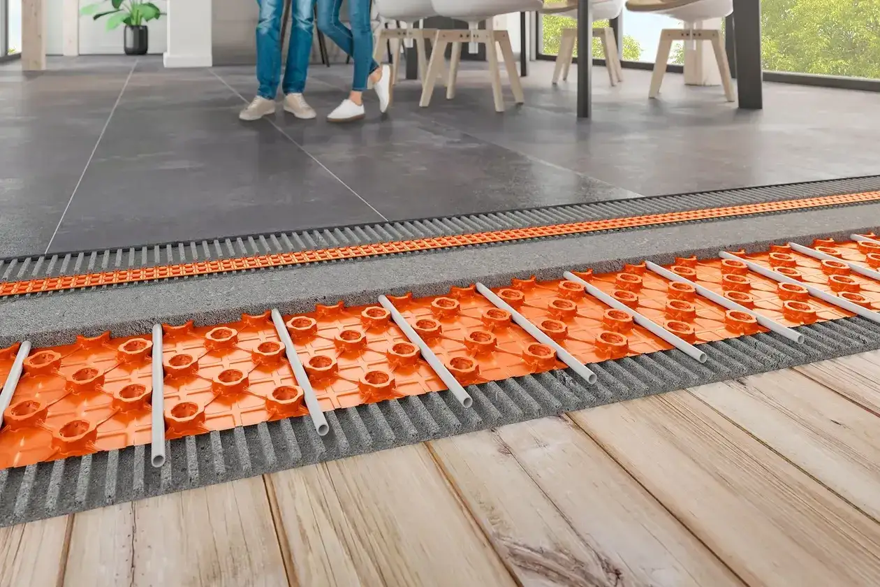 Vue en coupe d'un système de plancher chauffant, montrant les tuyaux orange sur une plaque alvéolée, sous un carrelage. Le cout plancher chauffant est optimisé.