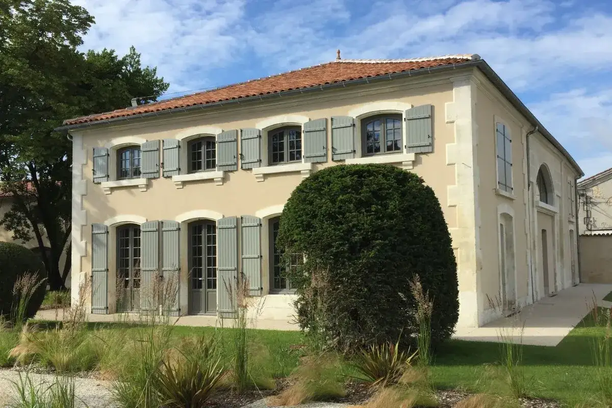 Belle maison aux volets gris, avec une grande fenêtre à l'anglaise au centre. Le jardin est luxuriant.