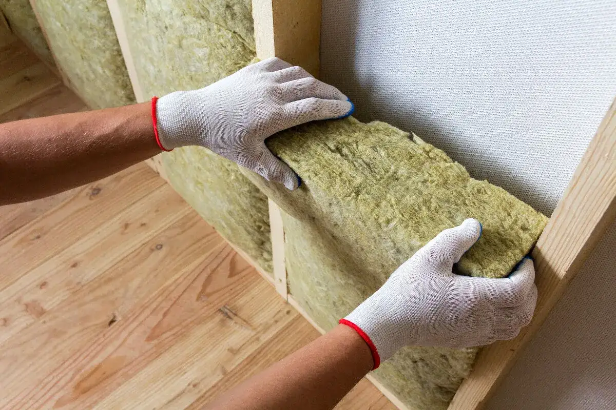 Mains gantées insèrent un panneau d'isolation minérale dans une structure en bois. L'isolation est essentielle pour le confort et l'efficacité énergétique.