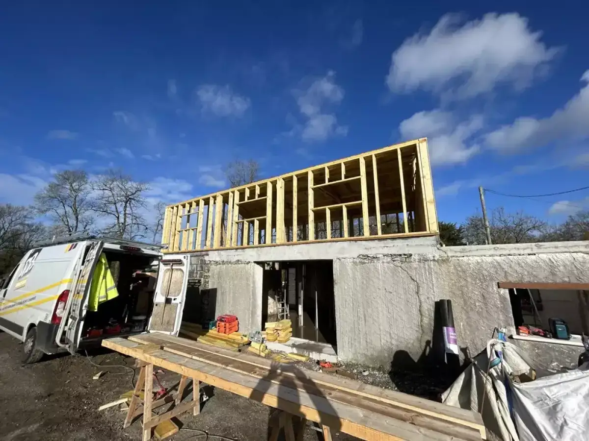 Chantier d'une extension bois, structure en bois visible sur un bâtiment en cours de rénovation. Un fourgon de chantier est garé à proximité.