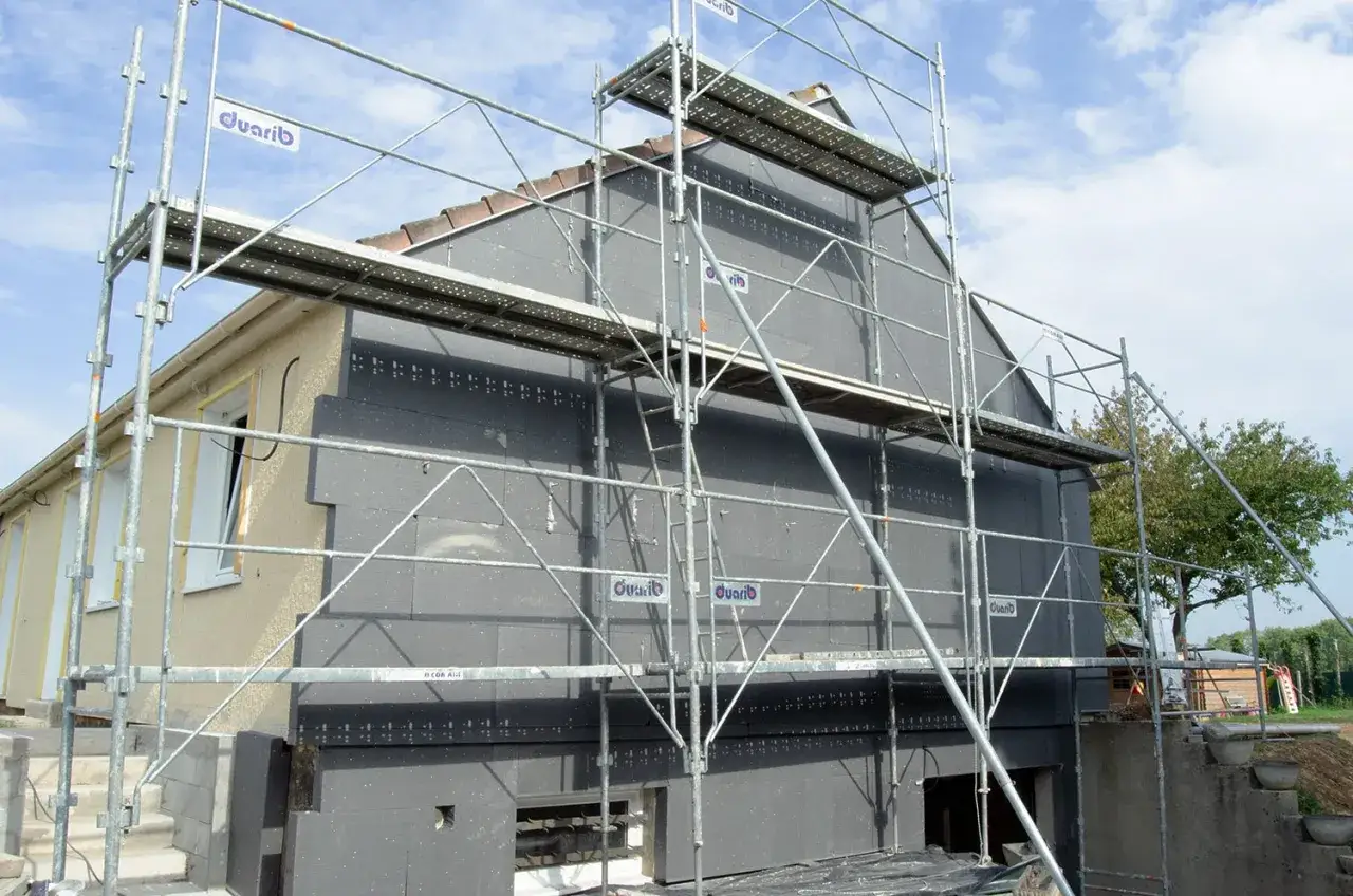 Chantier de rénovation : échafaudage autour d'une maison en cours d'isolation par l'extérieur avec des panneaux gris.
