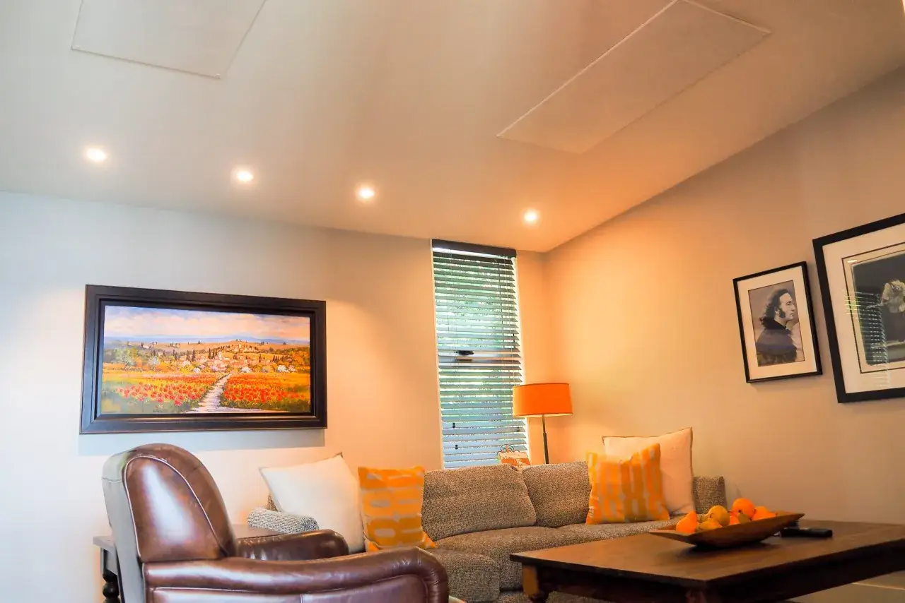 Salon confortable avec un tableau de paysage, un fauteuil en cuir et un canapé. Un panneau discret au plafond suggère un système de chauffage infrarouge.